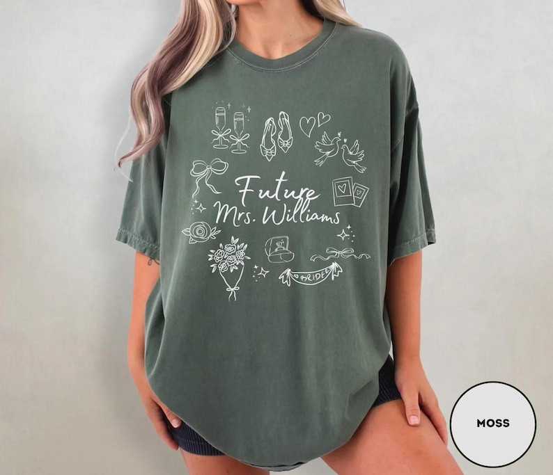 May include: Moss green t-shirt with a white graphic design. The design includes champagne glasses, shoes, doves, hearts, and the text "Future Mrs. Williams". A bouquet of flowers, a ring, and a banner that says "Bride" are also included.