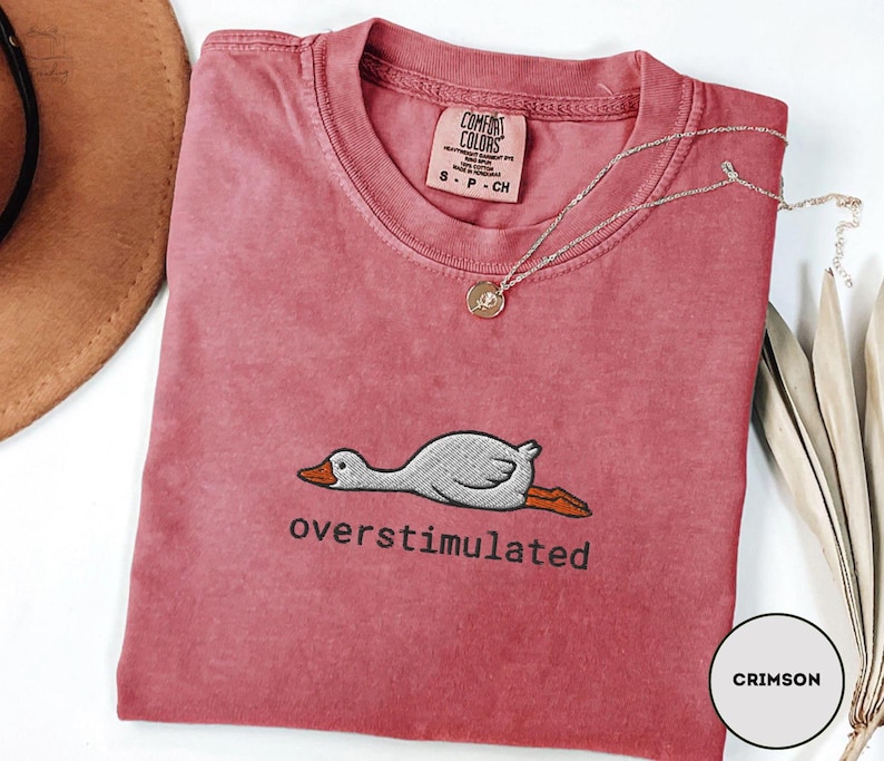 May include: Crimson-coloured t-shirt with a crew neck and short sleeves. The shirt features an embroidered graphic of a relaxed duck and the word "overstimulated" in black. A gold necklace with a pendant is draped on the shirt.