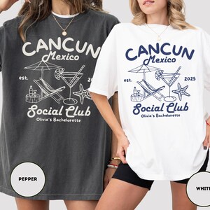 May include: Two oversized t-shirts with a graphic design featuring a beach scene with a cocktail, lounge chair, and palm tree. The text on the shirts reads "CANCUN Mexico Social Club Olivia's Bachelorette" with the year "2025" on the white shirt. The shirts are shown on models wearing black shorts. The shirt on the left is a dark gray color and the shirt on the right is white.