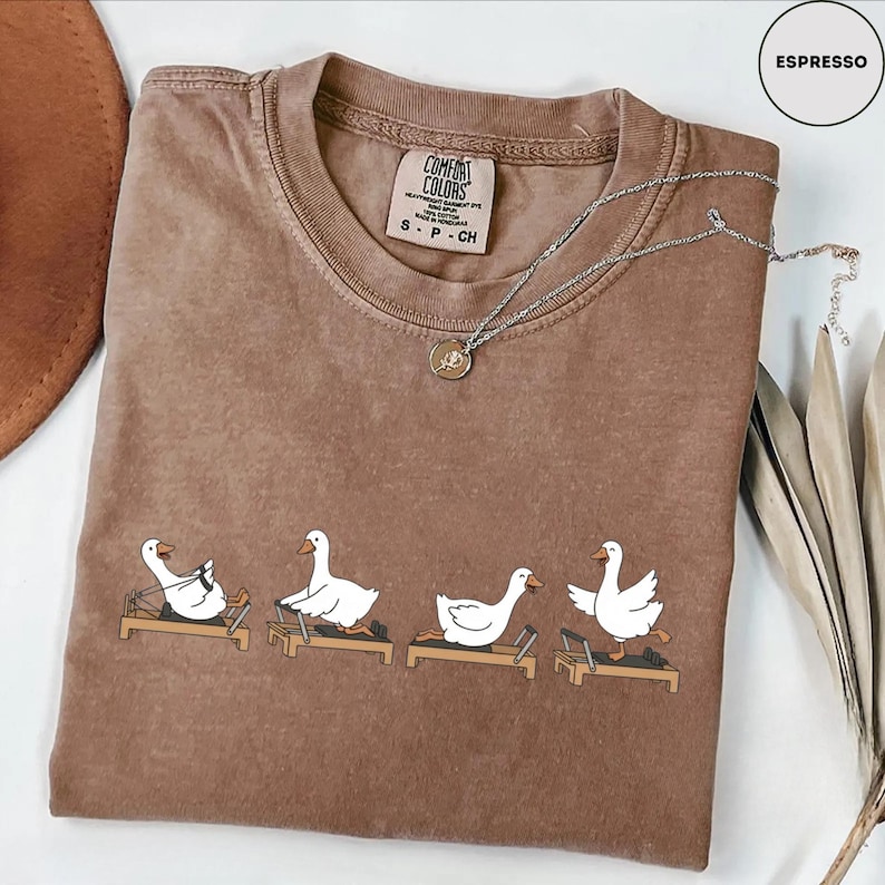 Puede incluir: Camiseta Comfort Colors color espresso con cuello redondo y mangas cortas. La camiseta presenta un gr&aacute;fico de gansos blancos de dibujos animados haciendo ejercicio en m&aacute;quinas de Pilates. Un collar plateado con un colgante est&aacute; drapeado sobre la camiseta.