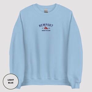 May include: Pale blue sweatshirt featuring the word "Newport" in navy blue, with a red lobster graphic and the text "16 / 39 Rhode Island" beneath. The sweatshirt is a solid pale blue colour.