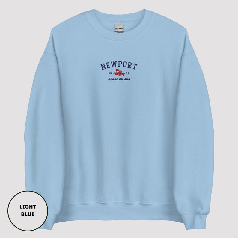 May include: Pale blue sweatshirt featuring the word "Newport" in navy blue, with a red lobster graphic and the text "16 / 39 Rhode Island" beneath. The sweatshirt is a solid pale blue colour.