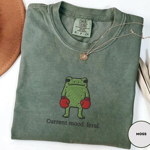 May include: Moss green t-shirt with a crew neck, featuring an embroidered green frog wearing red boxing gloves. The text "Current mood: feral." is below the frog. A gold necklace with a pendant is on the shirt.