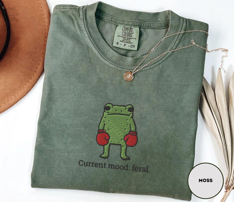 May include: Moss green t-shirt with a crew neck, featuring an embroidered green frog wearing red boxing gloves. The text "Current mood: feral." is below the frog. A gold necklace with a pendant is on the shirt.