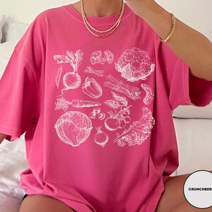 May include: A vibrant pink t-shirt featuring a detailed white illustration of various vegetables, including a beet, carrot, and cauliflower. The design is centered on the shirt, creating a bold statement. The shirt is made of a soft material.