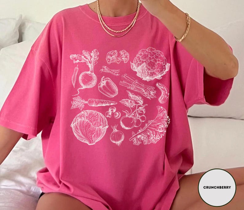 May include: A vibrant pink t-shirt featuring a detailed white illustration of various vegetables, including a beet, carrot, and cauliflower. The design is centered on the shirt, creating a bold statement. The shirt is made of a soft material.