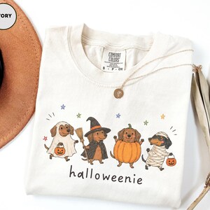 Op de afbeelding: Een ivoorkleurig t-shirt met een afbeelding van vier teckels in Halloween-kostuums: een spook, een heks met een bezem, een pompoen en een mummie. Elke hond draagt een pompoenemmer. De tekst "halloweenie" staat onder de honden gedrukt.