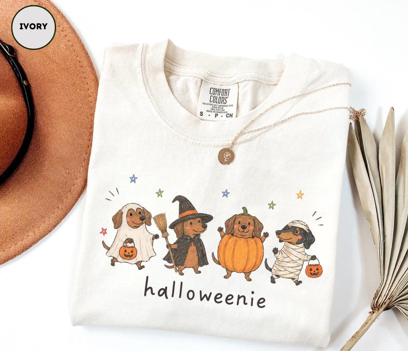 Op de afbeelding: Een ivoorkleurig t-shirt met een afbeelding van vier teckels in Halloween-kostuums: een spook, een heks met een bezem, een pompoen en een mummie. Elke hond draagt een pompoenemmer. De tekst "halloweenie" staat onder de honden gedrukt.