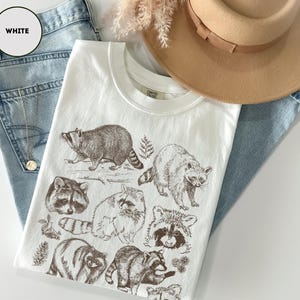 Op de afbeelding: Wit t-shirt met bruine inkt illustraties van wasberen. Het shirt wordt gecombineerd met lichtblauwe jeans en een bruine hoed. Het label van het shirt is zichtbaar. Er is ook een gouden ketting aanwezig.