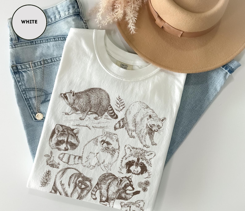 Op de afbeelding: Wit t-shirt met bruine inkt illustraties van wasberen. Het shirt wordt gecombineerd met lichtblauwe jeans en een bruine hoed. Het label van het shirt is zichtbaar. Er is ook een gouden ketting aanwezig.