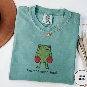 May include: A light green t-shirt with a cartoon frog wearing red boxing gloves. The text "Current mood: feral" is below the frog. A gold necklace is draped across the shirt.