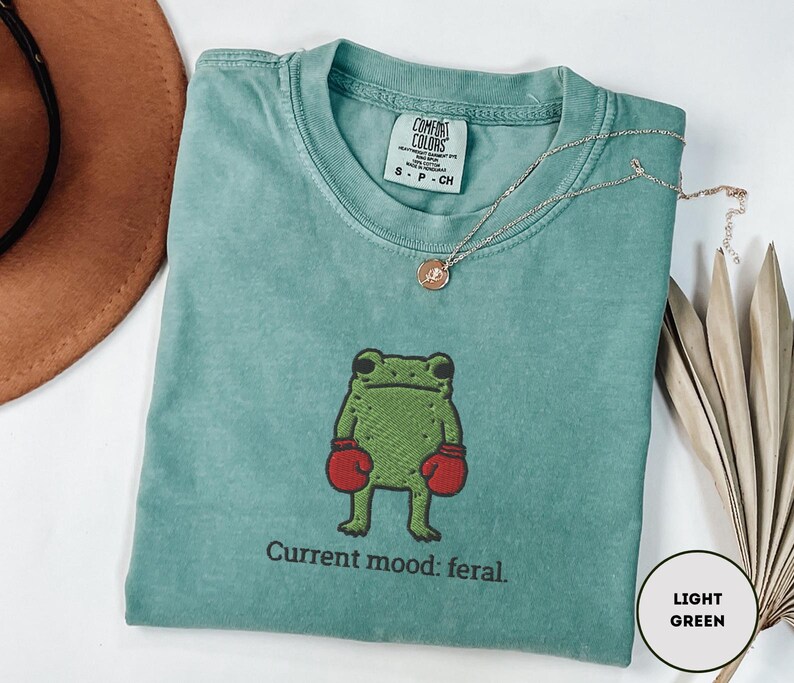 May include: A light green t-shirt with a cartoon frog wearing red boxing gloves. The text "Current mood: feral" is below the frog. A gold necklace is draped across the shirt.