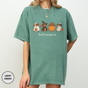 Op de afbeelding: Een lichtgroen T-shirt met korte mouwen en een afbeelding van vier teckels in Halloween-kostuums, waaronder een spook, een heks, een pompoen en een mummie. De tekst "halloweenie" is in het wit onder de honden gedrukt, samen met kleine sterretjes.