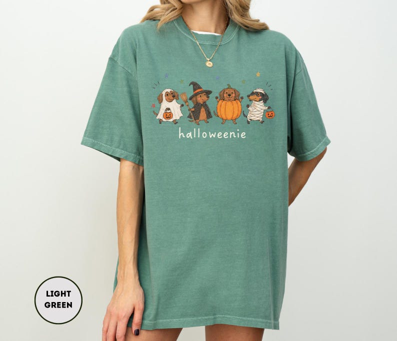 Op de afbeelding: Een lichtgroen T-shirt met korte mouwen en een afbeelding van vier teckels in Halloween-kostuums, waaronder een spook, een heks, een pompoen en een mummie. De tekst "halloweenie" is in het wit onder de honden gedrukt, samen met kleine sterretjes.