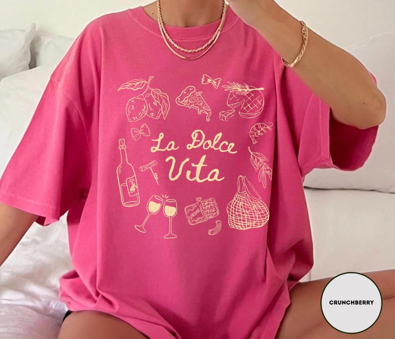 May include: A hot pink t-shirt with a cream-colored graphic design featuring Italian food illustrations and the text "La Dolce Vita". The design includes lemons, pizza, wine, and other Italian food items.