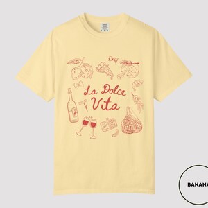 May include: Banana-colored t-shirt with a red illustration of Italian food and drink, including wine glasses, a bottle, and pizza. The words "La Dolce Vita" are written in red script. The shirt is a casual, short-sleeved style.