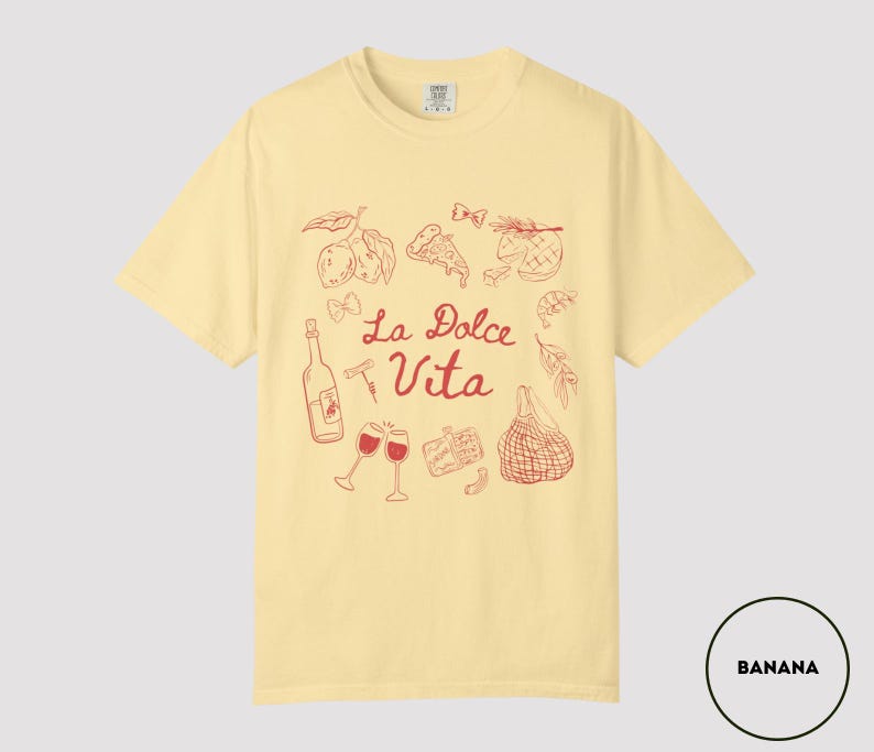 May include: Banana-colored t-shirt with a red illustration of Italian food and drink, including wine glasses, a bottle, and pizza. The words "La Dolce Vita" are written in red script. The shirt is a casual, short-sleeved style.