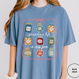 Peut inclure: T-shirt bleu jean avec un motif de bocaux de confiture contenant diff&eacute;rents fruits et le texte "grandma life is my jam". Le motif est encadr&eacute; par un n&oelig;ud et une ligne roses.