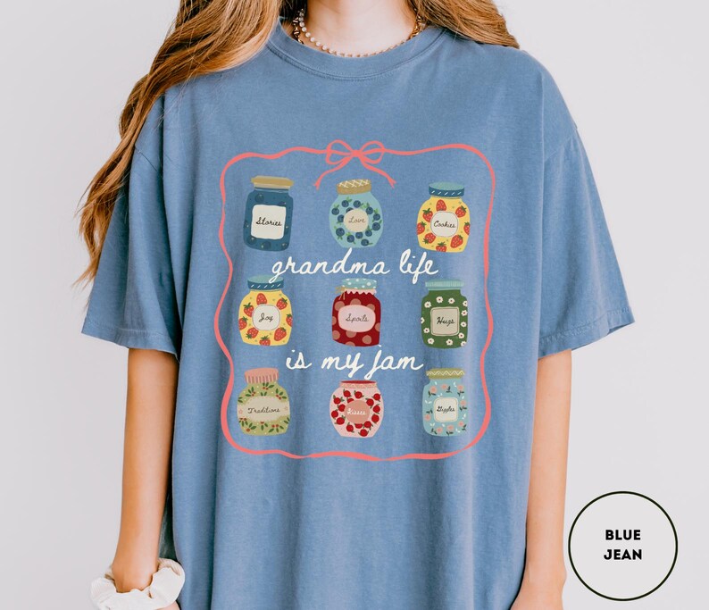 Peut inclure: T-shirt bleu jean avec un motif de bocaux de confiture contenant diff&eacute;rents fruits et le texte "grandma life is my jam". Le motif est encadr&eacute; par un n&oelig;ud et une ligne roses.