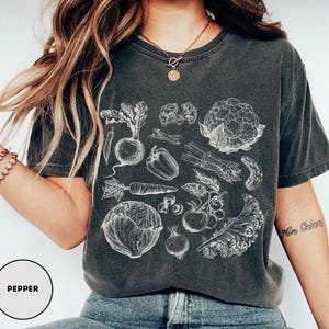 May include: Dark gray t-shirt featuring a detailed white illustration of various vegetables, including a carrot, pepper, and cauliflower. The word "PEPPER" is printed in a circle at the bottom left.