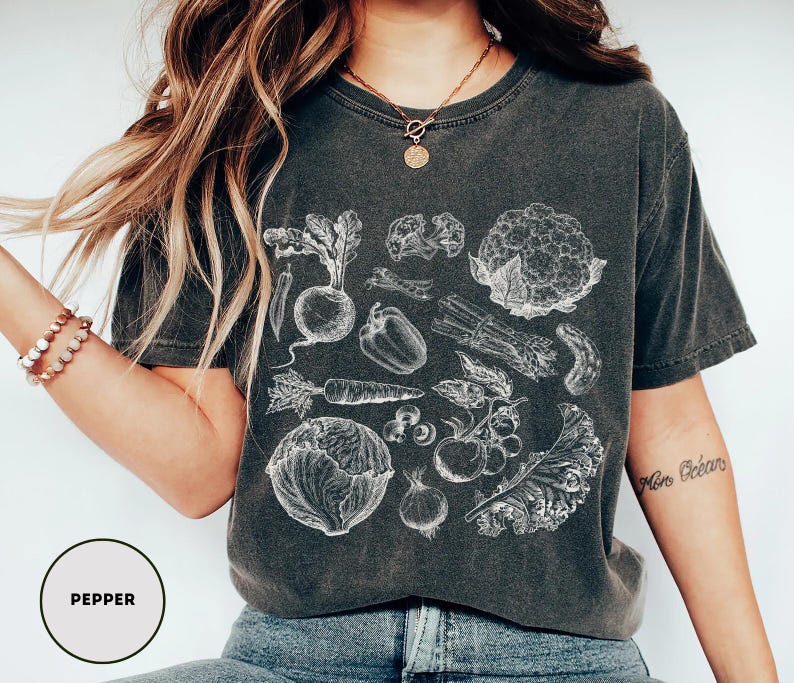 May include: Dark gray t-shirt featuring a detailed white illustration of various vegetables, including a carrot, pepper, and cauliflower. The word "PEPPER" is printed in a circle at the bottom left.