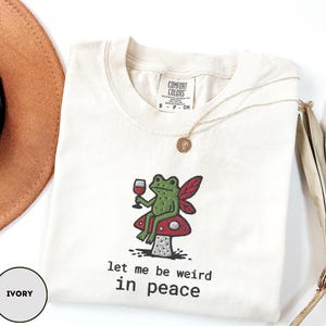 May include: Ivory-colored t-shirt with a whimsical frog design. The green frog, holding a wine glass, sits on a red mushroom with a white polka dot pattern. The text "let me be weird in peace" is below the frog. A gold necklace is draped on the shirt.
