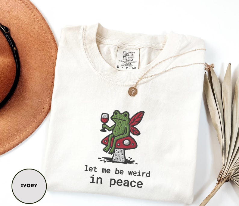 May include: Ivory-colored t-shirt with a whimsical frog design. The green frog, holding a wine glass, sits on a red mushroom with a white polka dot pattern. The text "let me be weird in peace" is below the frog. A gold necklace is draped on the shirt.