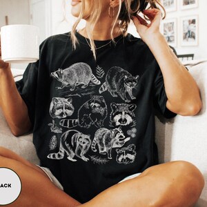 Op de afbeelding: Zwart T-shirt met een witte illustratie van wasberen in verschillende poses. Het ontwerp toont gedetailleerde tekeningen van wasberen, wat een op de natuur ge&iuml;nspireerde esthetiek cre&euml;ert. Het shirt is een comfortabel, casual kledingstuk.
