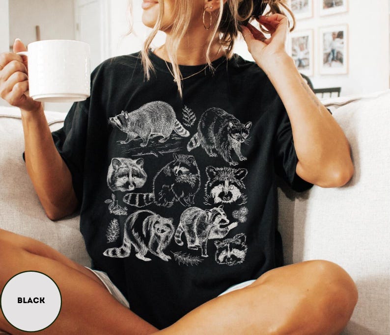 Op de afbeelding: Zwart T-shirt met een witte illustratie van wasberen in verschillende poses. Het ontwerp toont gedetailleerde tekeningen van wasberen, wat een op de natuur ge&iuml;nspireerde esthetiek cre&euml;ert. Het shirt is een comfortabel, casual kledingstuk.