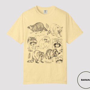 Op de afbeelding: Bananegele t-shirt met een reeks gedetailleerde wasbeer illustraties in zwarte inkt. Het ontwerp bevat verschillende poses en close-ups van de dieren. Het shirt heeft een klassieke ronde hals en korte mouwen.