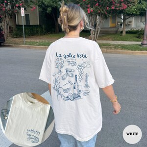 Puede incluir: Camiseta blanca con un estampado gr&aacute;fico azul de una botella de vino, uvas, ostras y una langosta. El texto "La Dolce Vita" est&aacute; impreso en la camiseta.
