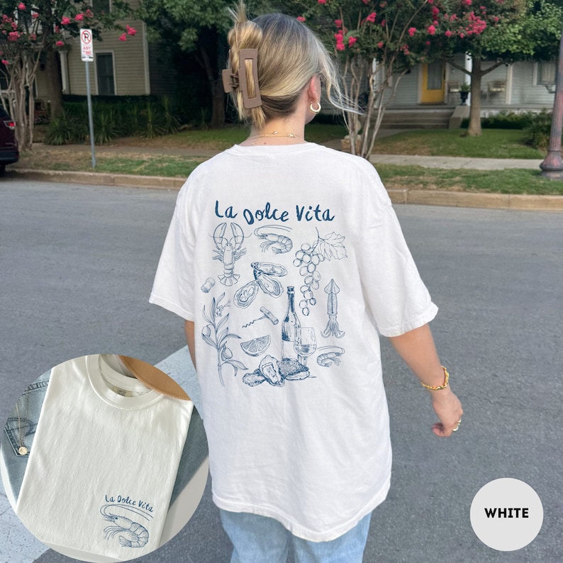 Puede incluir: Camiseta blanca con un estampado gr&aacute;fico azul de una botella de vino, uvas, ostras y una langosta. El texto "La Dolce Vita" est&aacute; impreso en la camiseta.