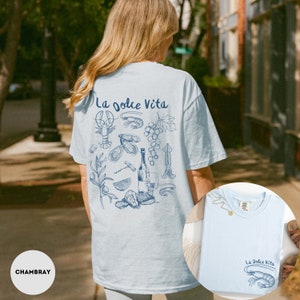 Puede incluir: Una camiseta azul claro con el texto "La Dolce Vita" impreso en la espalda. El dise&ntilde;o presenta una variedad de ilustraciones, incluyendo una langosta, uvas, vino y ostras.