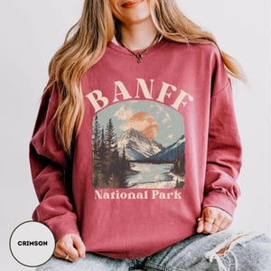 May include: A crimson red sweatshirt with a vintage-style graphic of a mountain range and a river, with the text "Banff National Park" printed below the image.