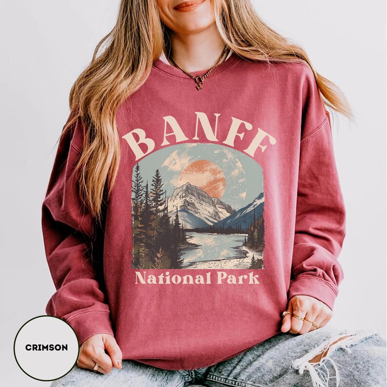 May include: A crimson red sweatshirt with a vintage-style graphic of a mountain range and a river, with the text "Banff National Park" printed below the image.
