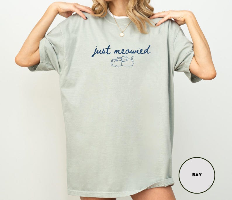 May include: A light green t-shirt with the text "just meowied" in navy blue script above a simple cat illustration. The shirt has short sleeves and a relaxed fit.