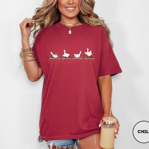 Puede incluir: Camiseta burdeos con un dise&ntilde;o caprichoso de cuatro gansos blancos en posturas de yoga sobre esterillas marrones. La camiseta tiene un corte holgado y de gran tama&ntilde;o. El color es un tono rojo intenso.
