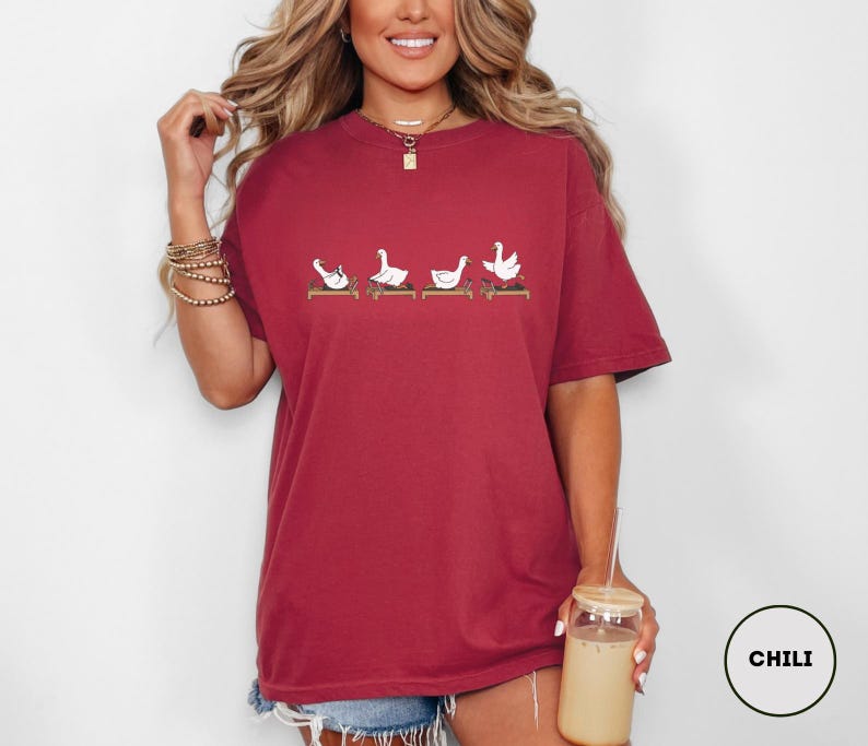 Puede incluir: Camiseta burdeos con un dise&ntilde;o caprichoso de cuatro gansos blancos en posturas de yoga sobre esterillas marrones. La camiseta tiene un corte holgado y de gran tama&ntilde;o. El color es un tono rojo intenso.