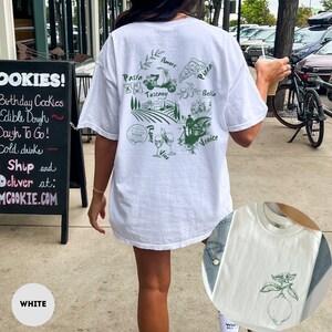 May include: White t-shirt with a green graphic of Italian imagery, including a scooter, pasta, pizza, and the words "Amore," "Tuscany," "Bella," and "Venice." The shirt also features a lemon graphic. The text "ROME" is also visible.
