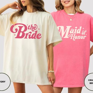 May include: Two oversized t-shirts. One is ivory with the text "the Bride" in pink script. The other is pink with the text "Maid of Honor" in white script. Both shirts have short sleeves and a crew neck.