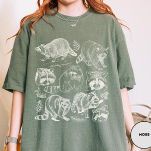 Op de afbeelding: Mosgroen T-shirt met een witte illustratie van wasberen in verschillende poses. Het ontwerp bevat gedetailleerde tekeningen van de dieren, sommige met bladeren. Het shirt heeft een relaxte pasvorm.