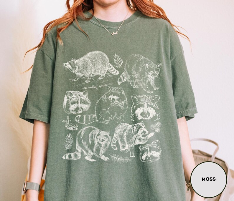 Op de afbeelding: Mosgroen T-shirt met een witte illustratie van wasberen in verschillende poses. Het ontwerp bevat gedetailleerde tekeningen van de dieren, sommige met bladeren. Het shirt heeft een relaxte pasvorm.