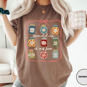 May include: Espresso-colored t-shirt featuring a colorful graphic of jam jars with the text "neonatal icu is my jam." The jars are filled with various fruits and labeled with text. The shirt is a casual, comfortable style.