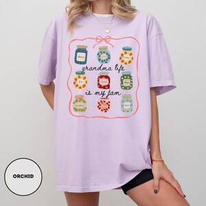 Peut inclure: T-shirt lilas clair avec un motif de bocaux de confiture color&eacute;s et l'inscription "grandma life is my jam". Le motif est encadr&eacute; d'une bordure rose. Le t-shirt est ample et confortable.