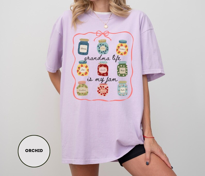 Peut inclure: T-shirt lilas clair avec un motif de bocaux de confiture color&eacute;s et l'inscription "grandma life is my jam". Le motif est encadr&eacute; d'une bordure rose. Le t-shirt est ample et confortable.