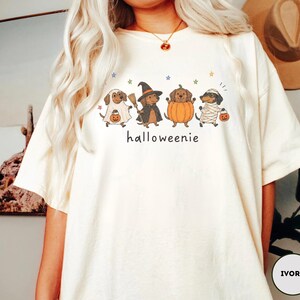 Op de afbeelding: Een ivoorkleurig t-shirt met een afbeelding van vier teckels in Halloween-kostuums, waaronder een spook, een heks, een pompoen en een mummie, met de tekst "halloweenie" eronder gedrukt en kleine gekleurde sterretjes eromheen verspreid.