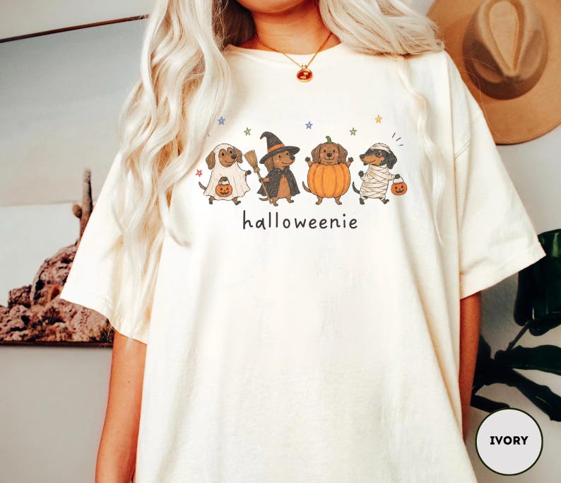 Op de afbeelding: Een ivoorkleurig t-shirt met een afbeelding van vier teckels in Halloween-kostuums, waaronder een spook, een heks, een pompoen en een mummie, met de tekst "halloweenie" eronder gedrukt en kleine gekleurde sterretjes eromheen verspreid.