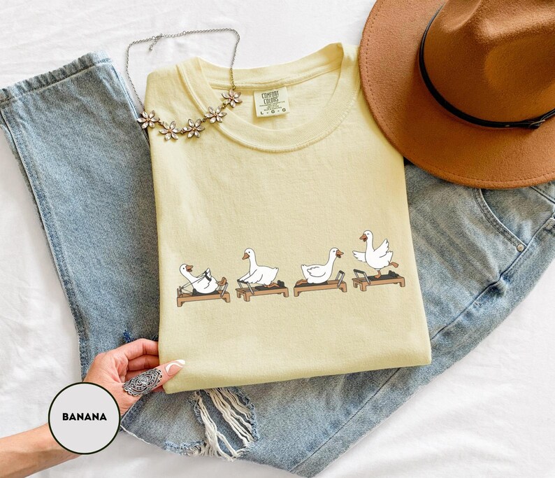 Puede incluir: Una camiseta amarilla clara con un gr&aacute;fico de cuatro gansos blancos haciendo ejercicio en cintas de correr. La camiseta est&aacute; doblada sobre unos vaqueros azules, con un sombrero marr&oacute;n y un collar floral. La palabra "BANANA" est&aacute; en un c&iacute;rculo en la esquina inferior izquierda.