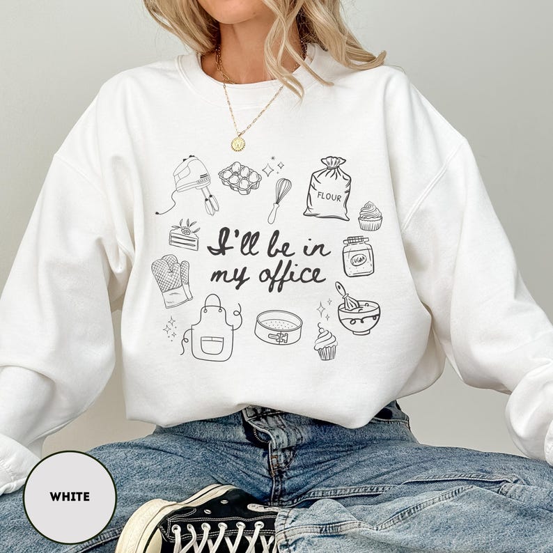 May include: White sweatshirt with a black line drawing of baking tools and the text "I'll be in my office." The design includes a mixer, whisk, apron, and cupcakes. The sweatshirt is a casual, comfortable style.