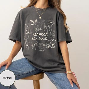 May include: Dark grey t-shirt with the text "respect the locals" and illustrations of various animals, including a wolf, deer, and bear. The shirt is worn by a person wearing blue jeans.